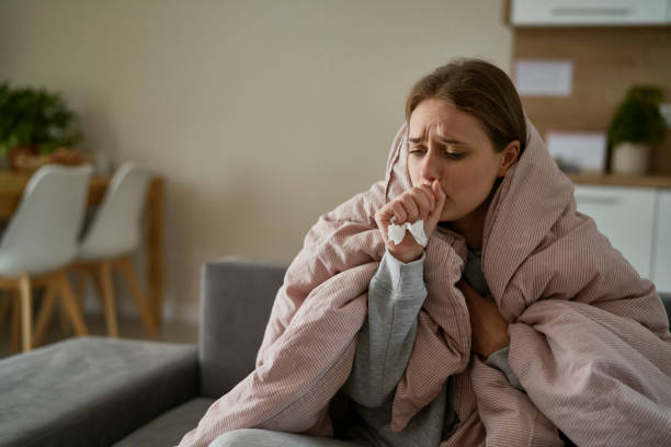 caucasian young woman coughing and sitting under the duvet at home